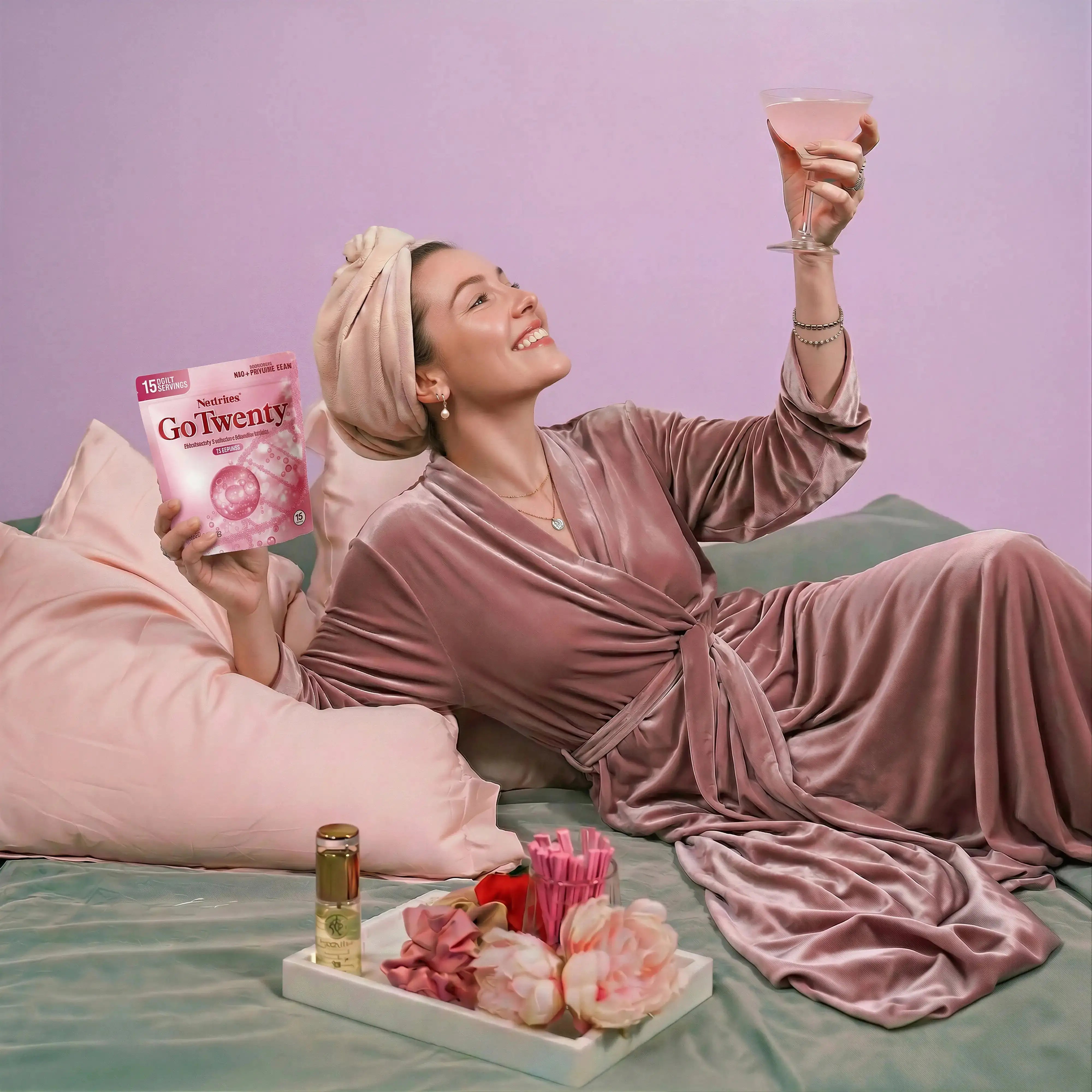 Woman in a pink robe holding a GoTwenties product and a glass, with flowers and a tray on a bed.